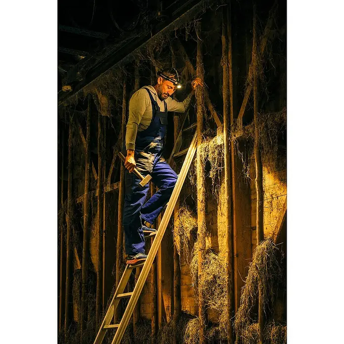 Ledlenser Stirnlampe H7R Work - 15 lm - 1.000 lm, IP67 - Ein Handwerker mit Stirnlampe, blauen Overalls und Hammer, steigt auf einer Leiter. Er inspiziert eine Wand mit Strohfüllung.