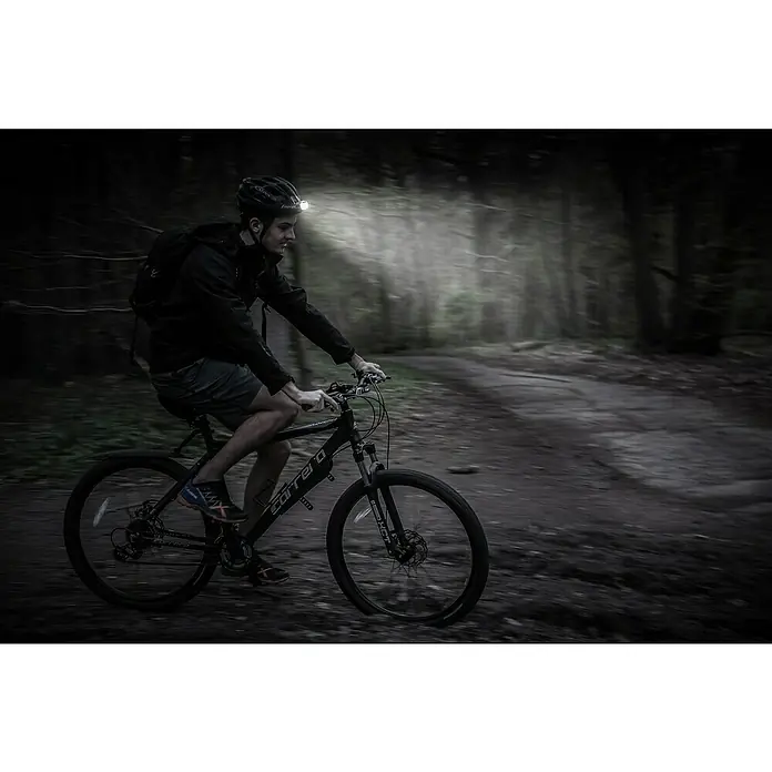 Energizer Linterna frontal deporte - 36 x 190 x 146 mm, 125 lm - Un hombre monta en una bicicleta de montaña Carrera en un sendero forestal oscuro, con un casco con una luz que ilumina el camino.