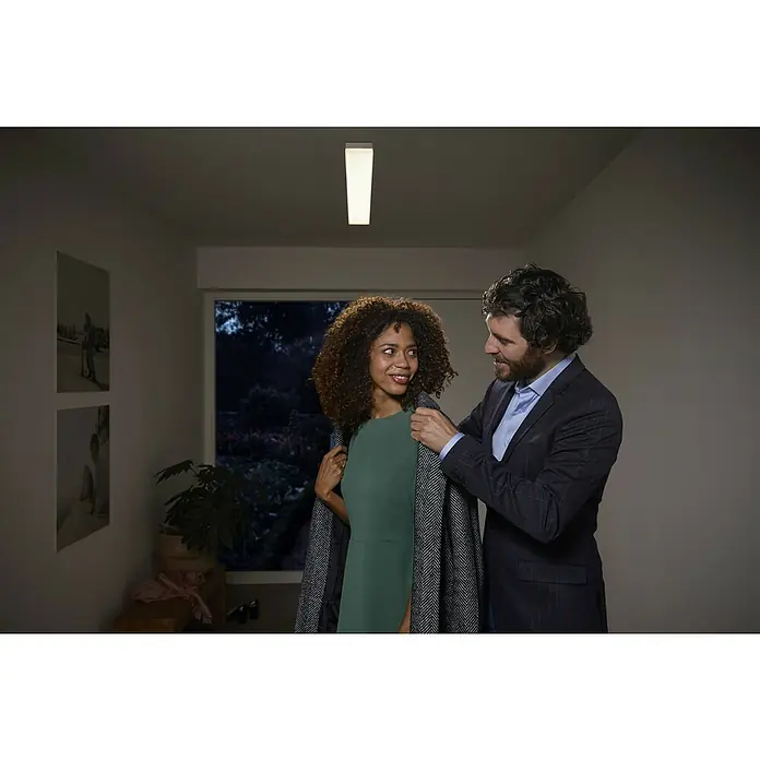 Ledvance Smart+ WiFi LED-Panel Planon Frameless - 28 W, L x B x H: 600 x 100 x 65 mm, Weiß - Eine moderne Deckenleuchte erhellt einen Flur, während ein Mann einer Frau einen Mantel umlegt, bevor sie das Haus verlassen.