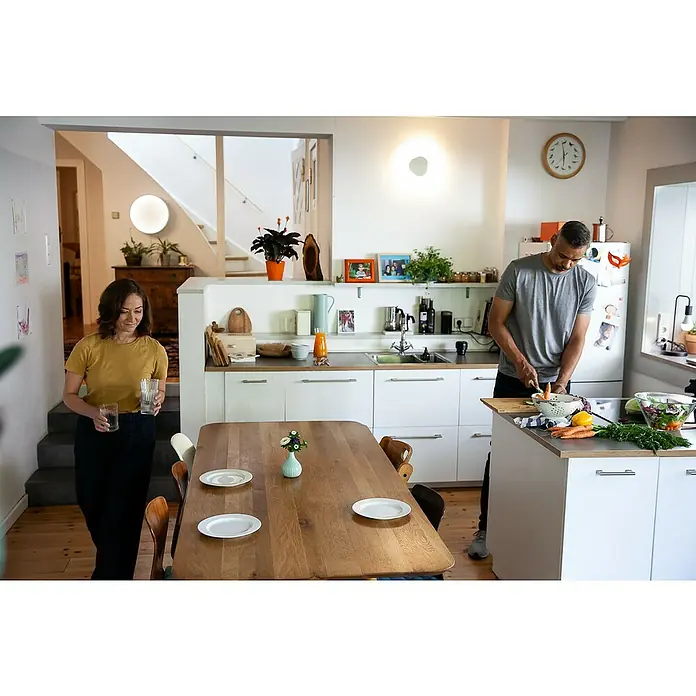 Ledvance Smart+ WiFi LED-Deckenleuchte rund Cylinder - 24 W, Ø x H: 450 x 134 mm, Weiß - Ein helles, modernes Esszimmer mit einem Holztisch, weißen Tellern, einer Vase und Menschen, die in der Küche beschäftigt sind.