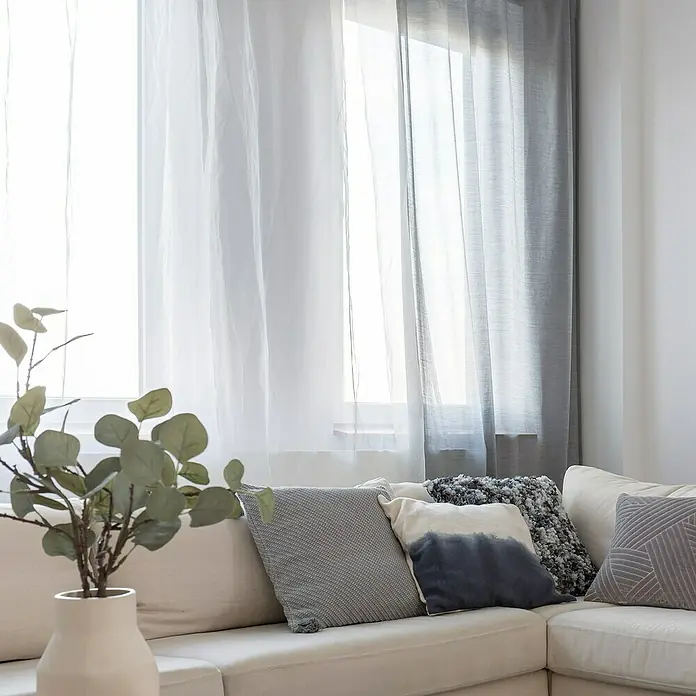 Cortina plisada con fruncido Linum - An x Al: 300 x 300 cm, Blanco, Materiales naturales - Rincón de sofá luminoso con cojines en tonos grises y azules, complementado con una rama decorativa sutil en un jarrón beige y cortinas translúcidas.