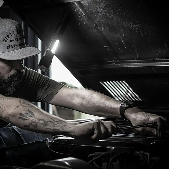 Nebo Tools Linterna Newton - 4,57 x 4,57 x 19,81 cm, 500 lm, IPX4 - Un mecánico trabaja bajo el capó de un coche con una luz de trabajo brillante, enfocándose en la inspección y reparación.