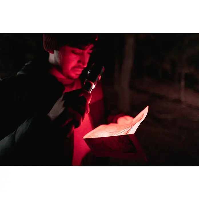 Nebo Tools Linterna Newton - 4,57 x 4,57 x 19,81 cm, 500 lm, IPX4 - Un hombre con gorro ilumina un mapa en la oscuridad con una linterna. La iluminación roja añade un ambiente misterioso a la imagen.