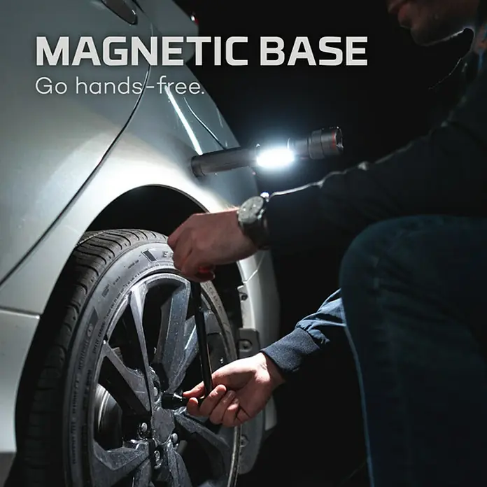 Nebo Tools Linterna Newton - 4,57 x 4,57 x 19,81 cm, 500 lm, IPX4 - La imagen muestra una luz de trabajo con base magnética adherida a un coche, mientras alguien trabaja en la rueda.