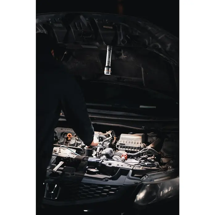 Nebo Tools Linterna táctil Newton - 3,81 x 4,78 x 17,78 cm, 1.000 lm, IP67 - Un mecánico se inclina sobre el compartimento del motor de un coche, usando una linterna para ayudar con las reparaciones.