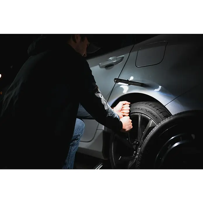 Nebo Tools Linterna Newton - 1,78 x 1,78 x 21,34 cm, 400 lm, IPX4 - Una persona monta un neumático en un coche plateado, con una luz de trabajo magnética para ayudar con la tarea y proporcionar una visibilidad clara.