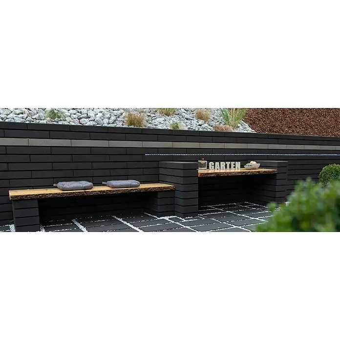 Christoph Mauerstein C-Line - Anthrazit, 50 x 25 x 10 cm, Beton - Gartenbereich mit schwarzer Mauer, Holzbank und Tisch. Die Mauer ist mit Pflanzen und einer Lichterkette dekoriert und der Boden mit Steinen gepflastert.