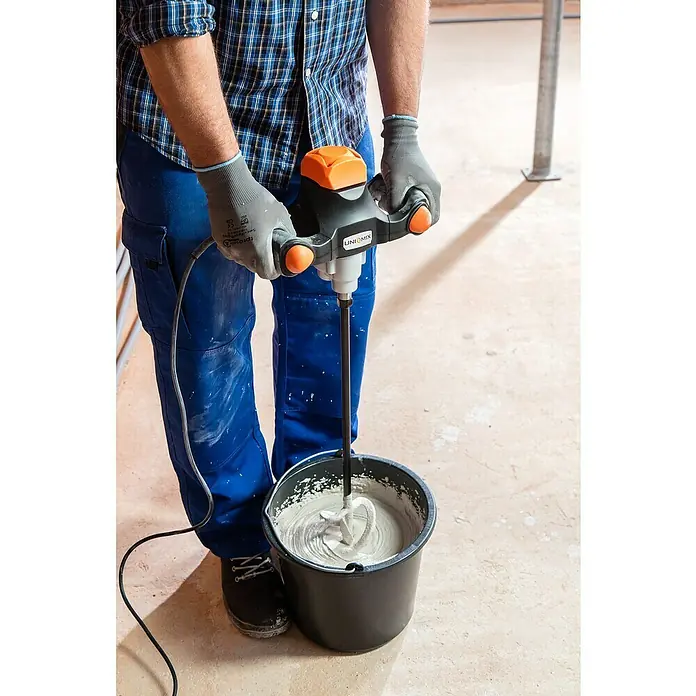 Collomix UniMix Handrührwerk 900 - Durchmesser Rührer: 120 mm, M 14, Mischvolumen: 40 l - Ein Bauarbeiter mischt Beton mit einem UNIQ-MIX Mörtelrührer, wobei die orangefarbenen Griffe hervorstechen, während er Schutzkleidung und Handschuhe trägt.