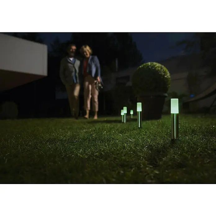 Ledvance Smart+ Erdspießleuchte Garten Pole RGBW Erweiterungs-Set - Farbe Gehäuse: Silber, RGBW, Mit Leuchtmittel - Nachtaufnahme von mehreren grünen Gartenleuchten, die einen Gartenweg beleuchten, mit einem Paar im Hintergrund.