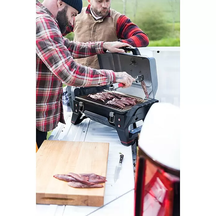 Char-Broil Gasgrill Grill2Go - Grillfläche (B x T): 44 x 28 cm, 2,7 kW, Anzahl Brenner: 2 - Ein Mann grillt Fleisch auf einem schwarzen tragbaren Grill im Freien, ein anderer Mann schaut zu. Rohe Fleischscheiben liegen auf einem Schneidebrett.