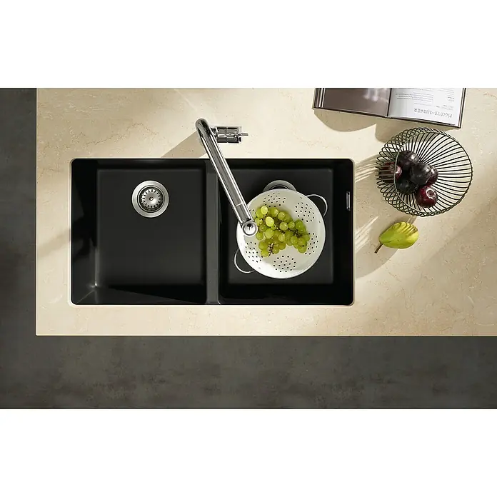 Hansgrohe Fregadero encastrable S510 - U770 2S - 45 x 82 cm, Piedra natural, Número de cubetas: 2 ud., Grafito, Resplandeciente - Un fregadero de cocina negro moderno con escurridor, un colador lleno de uvas verdes, al lado de un bol con ciruelas y una pera.