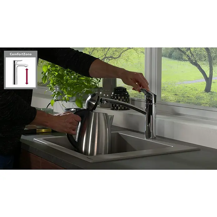 Hansgrohe Grifo de cocina MyCube M - Cromo, Brillante - Una persona llena un hervidor plateado en el grifo de un fregadero de cocina debajo de una ventana. El grifo tiene un diseño elegante y moderno.