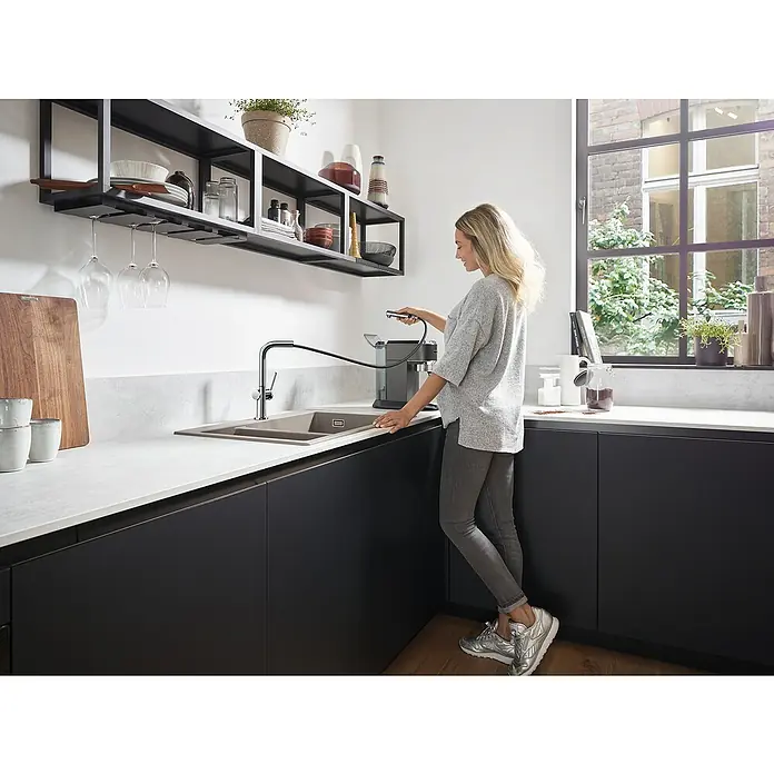 Hansgrohe Grifo de cocina Talis M54 - Cromo, Brillante - Cocina moderna con fregadero y grifo de acero inoxidable, mujer operando una máquina de café. La cocina cuenta con gabinetes negros y encimera clara.