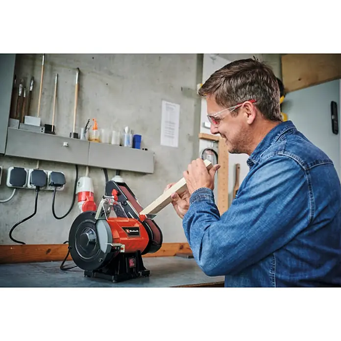 Einhell Esmeriladora doble eléctrica combinada TC-US 350 - 350 W, 2.980 r.p.m. - Un hombre en su taller usa una amoladora de banco Einhell para alisar un trozo de madera mientras usa gafas de seguridad para protección.