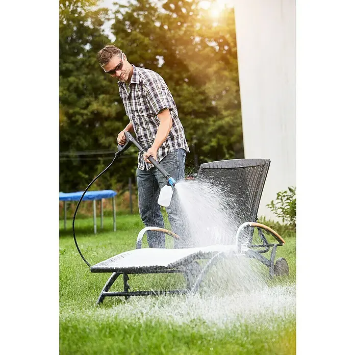 Nilfisk Detergente para plásticos Rattan & Plastic Cleaner - 2,5 l - Un hombre limpia una tumbona de patio gris con una hidrolavadora y espuma.