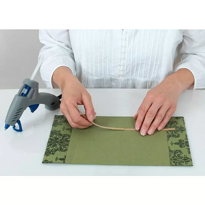 Dremel Barra de cola multiuso GG 11 - 11 mm, Específico para: Pistola termoadhesiva mod. 940, 12 ud. - Una persona usa una pistola de pegamento caliente Dremel 930 gris y azul para fijar una tira a tela verde.