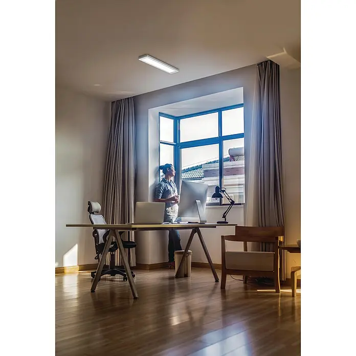 Ledvance LED-Deckenleuchte Office Line - 25 W, L x B x H: 61,5 x 13 x 2,5 cm, Weiß, Neutralweiß - Helles Büro mit einer Deckenleuchte, die für eine gute Ausleuchtung sorgt, während eine Person am Fenster steht und die Aussicht genießt.