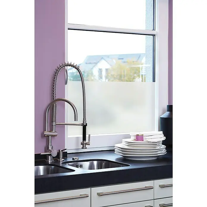 D-c-fix Static Premium Glasfolie Frost - 150 x 67,5 cm, Transluzent, Statisch haftend - Eine moderne Küchenspüle aus Edelstahl mit einem hohen, spiralförmigen Wasserhahn und Geschirr auf einer dunklen Arbeitsplatte, vor einem Fenster.