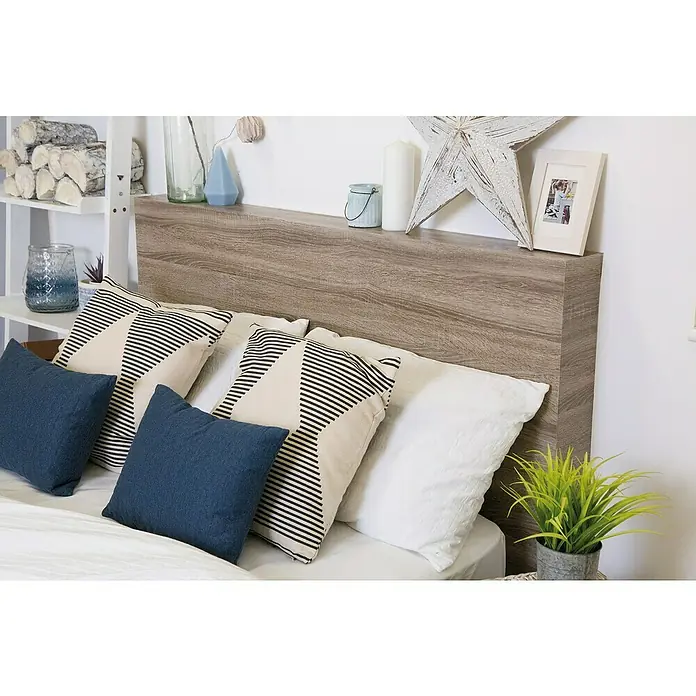 D-c-fix Holzoptikfolie Sonoma Eiche - 200 x 67,5 cm, Trüffel, Selbstklebend - Nahaufnahme eines Bettes mit einem Kopfteil aus Holz, blauen und gemusterten Kissen sowie einem Nachttisch mit Grünpflanzen.