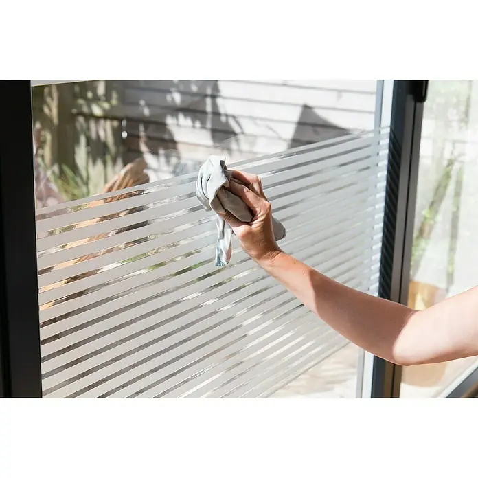 D-c-fix StaticStripes Glasfolie Clarity - 200 x 15 cm, Transluzent, Statisch haftend - Eine Person wischt mit einem grauen Tuch eine Fensterfolie mit horizontalen Streifen ab. Die Folie sorgt für Privatsphäre und Lichtdurchlässigkeit.