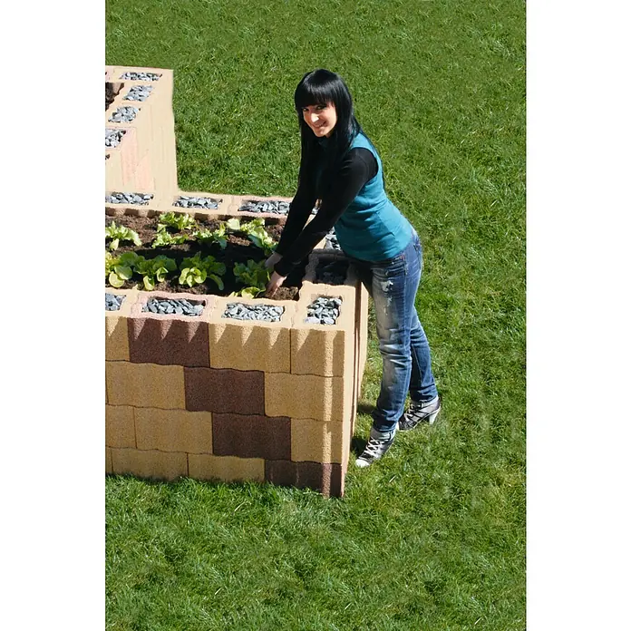 Christoph Pflanzstein Rasterstein - Mediterran, 30 x 20 x 20 cm, Beton - Frau beim Bepflanzen eines Hochbeets aus braunen und gelben Pflanzsteinen, gefüllt mit Erde, Steinen und Salatsetzlingen, auf einer grünen Wiese.