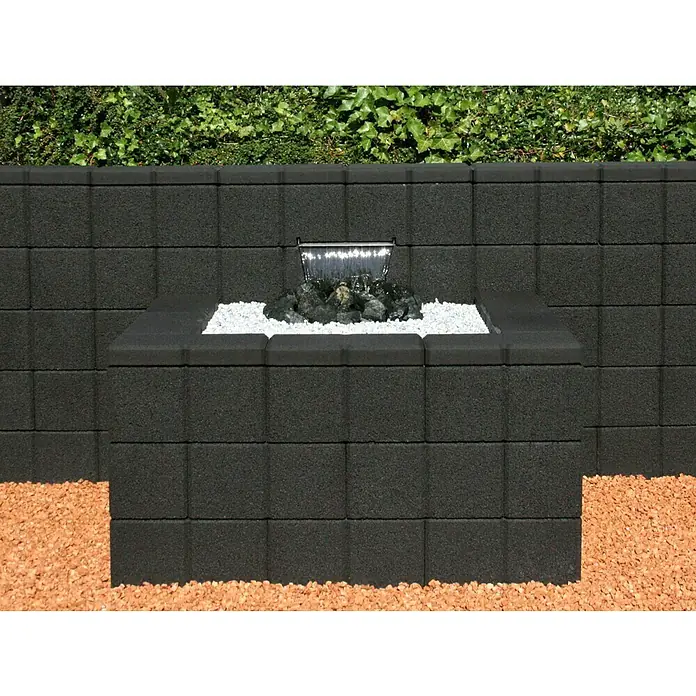 Christoph Gartenmauer Ambienta - 40 x 20 x 20 cm, Anthrazit, Beton - Nahaufnahme einer modernen schwarzen Brunnenwand mit Kieselsteinen und Wasserfall, umgeben von einem Hintergrund aus üppigem Grün.