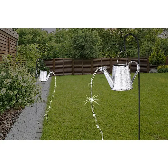 BAUHAUS Solar-Dekoleuchte Gießkanne auf Erdspitz - 0,06 W, L x B x H: 30 x 17 x 112 cm, Schwarz/Silbergrau, IP44 - Dekorative Gartenszene mit zwei beleuchteten, hängenden Gießkannen, die einen glitzernden Wassereffekt erzeugen und eine schöne Atmosphäre schaffen.