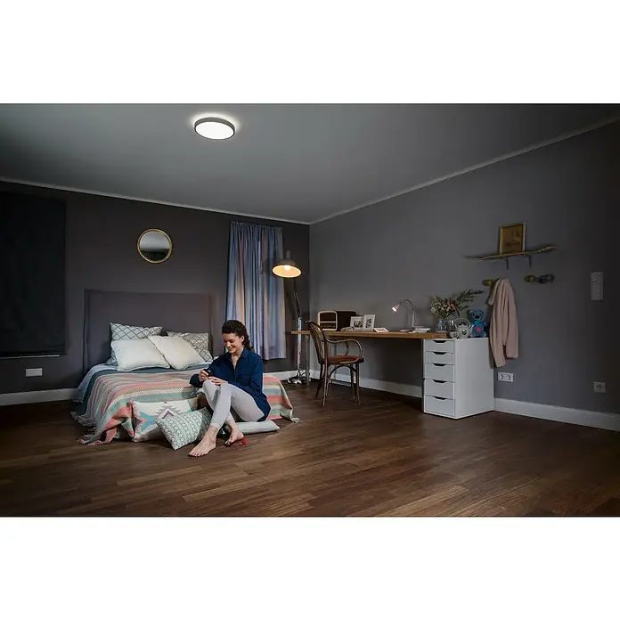 Ledvance LED-Deckenleuchte rund Silara - 16 W, Ø x H: 300 x 85 mm, Silber, Weiß, Neutralweiß - Ein modernes Schlafzimmer mit einer Deckenleuchte, einer Frau auf dem Bett, einem Schreibtisch und einem Bücherregal.