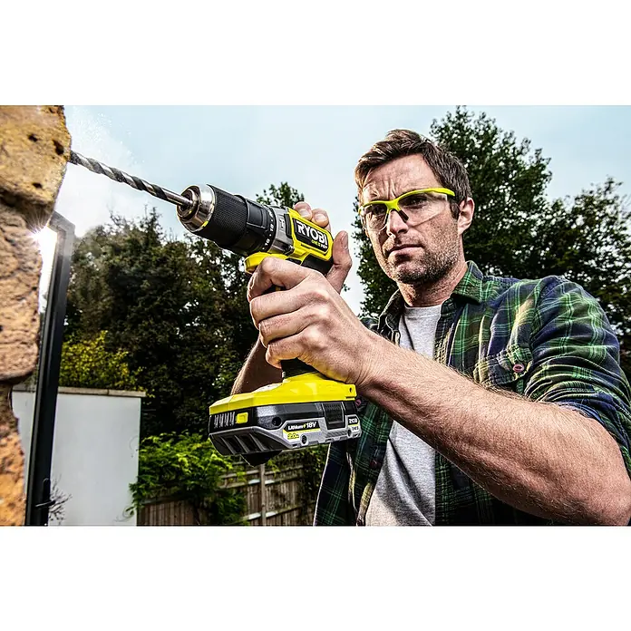 Ryobi ONE+HP Taladro atornillador percutor de batería RPD18C - 18 V, Sin batería, 0 Ah, 50 Nm - Un hombre perfora una pared con un taladro inalámbrico amarillo Ryobi para sujetar algo, usando gafas de seguridad.