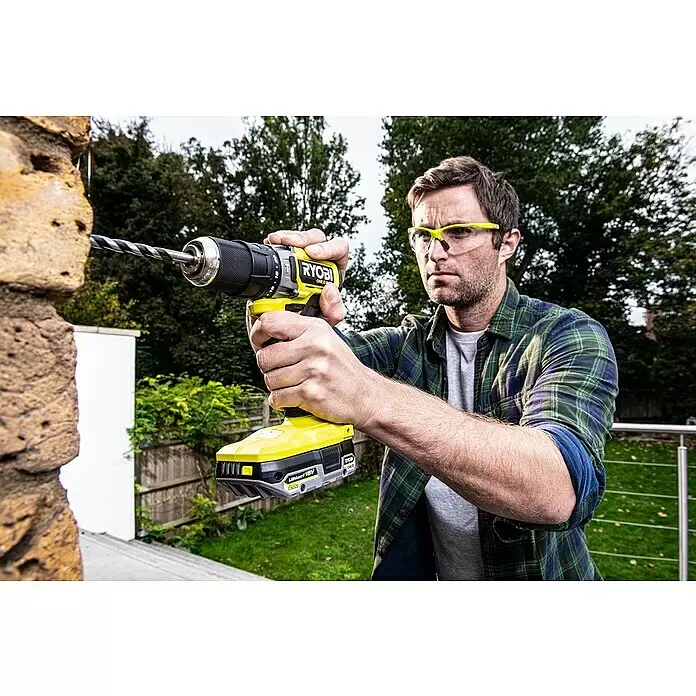 Ryobi ONE+HP Taladro atornillador percutor de batería RPD18C - 18 V, Sin batería, 0 Ah, 50 Nm - Hombre con gafas de seguridad usa un taladro inalámbrico Ryobi amarillo y negro para perforar una pared de piedra.