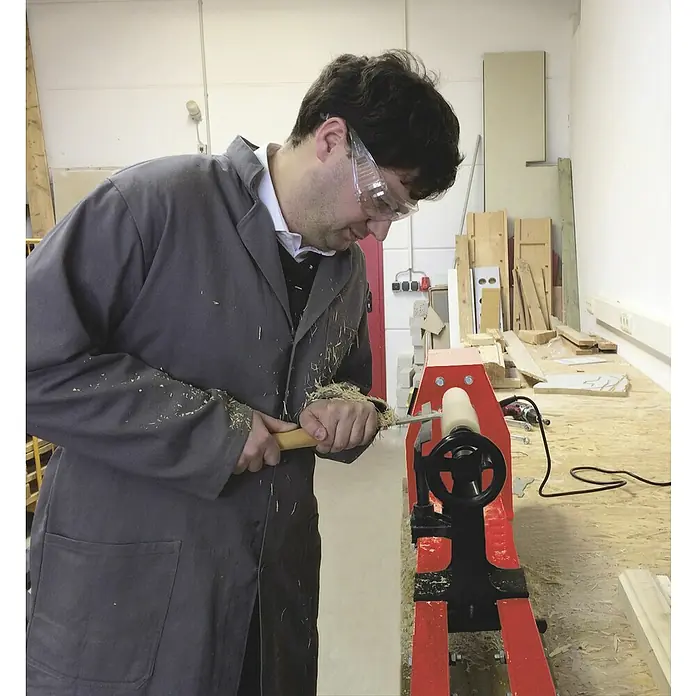 KWB Set de formones - 5 pzs. - Un hombre está trabajando en un torno de madera rojo, dando forma a una pieza con un cincel. Lleva gafas de seguridad y bata gris.