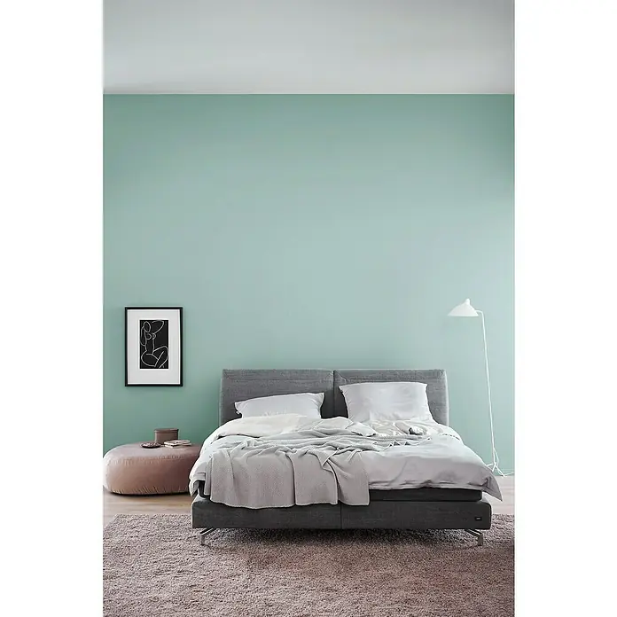 SCHÖNER WOHNEN-Farbe Wandfarbe Trendfarbe - Maui, Matt, 1 l, Konservierungsmittelfrei - Bett mit grauem Rahmen, weißer Bettwäsche und einer grauen Decke darauf. Ein minimalistisches Gemälde und eine Stehlampe schmücken den Raum.