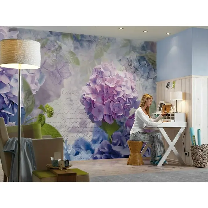 Komar Imagine Edition 3 - Stories Fototapete Otaksa - 8 -tlg., B x H: 368 x 254 cm, Papier - Eine Frau sitzt an einem Schreibtisch in einem Raum mit einer floralen Tapete. Der Raum ist hell und freundlich gestaltet.