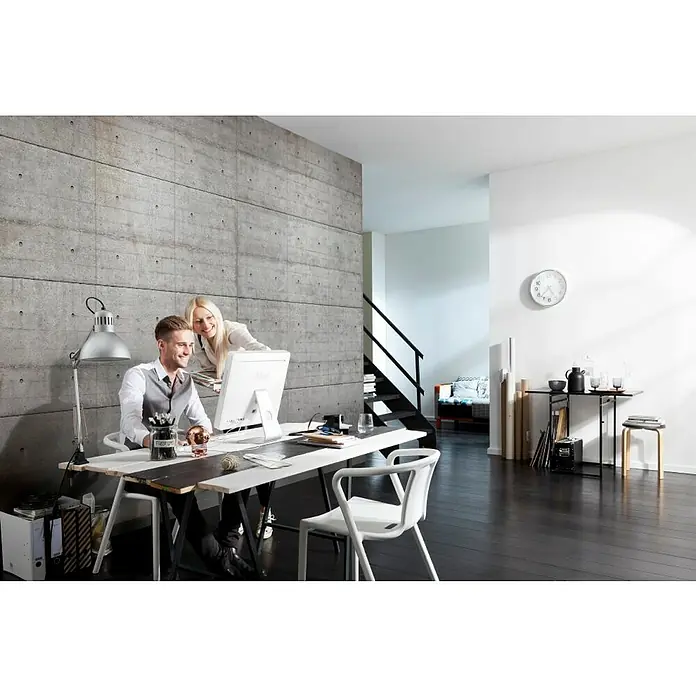 Komar Imagine Edition 3 - Stories Fototapete Concrete blocks - 8 -tlg., B x H: 245 x 368 cm, Papier - Modernes Büro Interieur mit Schreibtisch, Lampe und zwei Personen, die an einem Computer arbeiten, vor einer Betonwand.
