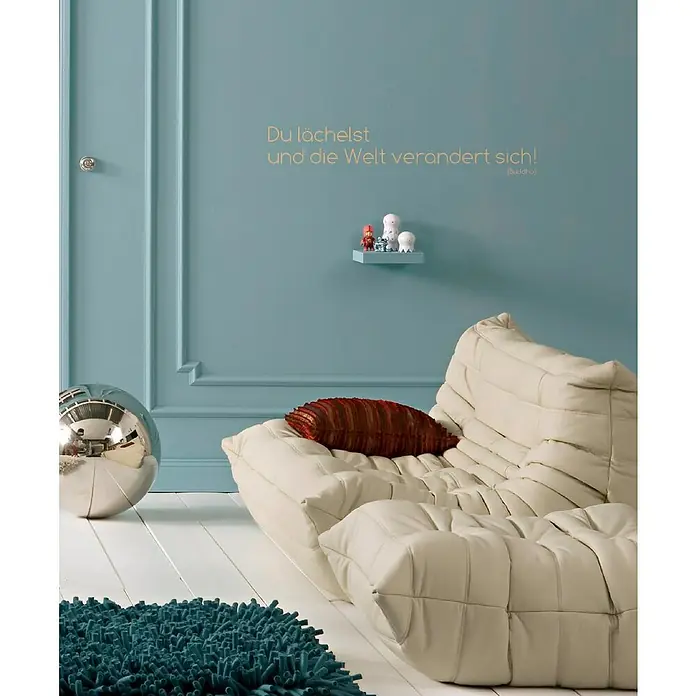 Komar Dekosticker Du Lächelst - 3 -tlg., 70 x 14 cm - Das Bild zeigt ein modernes Wohnzimmer mit einem beigen Sofa, einem blauen Teppich und einer silbernen Kugel.