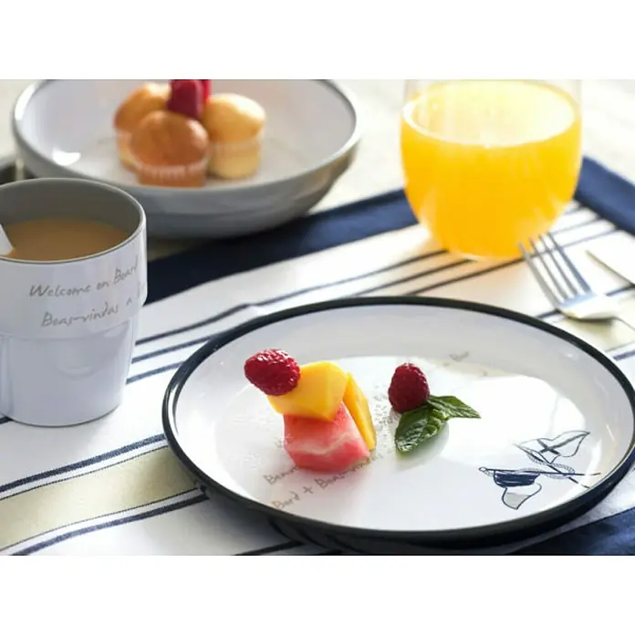Marine Business Welcome on Board Plato redondo - 6 ud., Diámetro: 21 cm - Bodegón de desayuno con fruta fresca, mini pasteles y zumo de naranja, presentado sobre un camino de mesa a rayas.