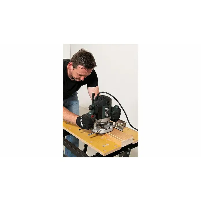 PowerPlus Fresadora eléctrica POWP6050 - 2.100 W, 8.000 r.p.m. - 23.500 r.p.m., Diámetro del portaherramientas: 12 mm - Un hombre utiliza una fresadora para cortar un trozo de madera, centrándose en la herramienta y su trabajo de precisión.