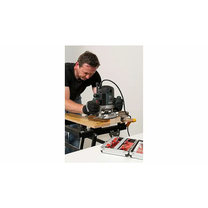 PowerPlus Fresadora eléctrica POWP6050 - 2.100 W, 8.000 r.p.m. - 23.500 r.p.m., Diámetro del portaherramientas: 12 mm - Un hombre utiliza una enrutadora de madera sobre un trozo de madera. La caja de herramientas con brocas de enrutador también está en la imagen.
