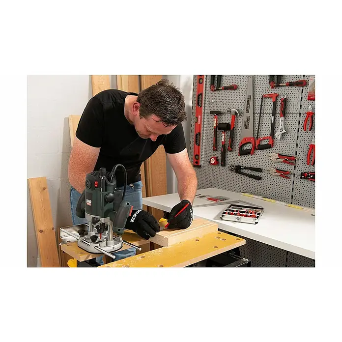 PowerPlus Fresadora eléctrica POWP6050 - 2.100 W, 8.000 r.p.m. - 23.500 r.p.m., Diámetro del portaherramientas: 12 mm - Un hombre utiliza una fresadora en una pieza de madera en su taller, con herramientas colgadas en la pared detrás de él.
