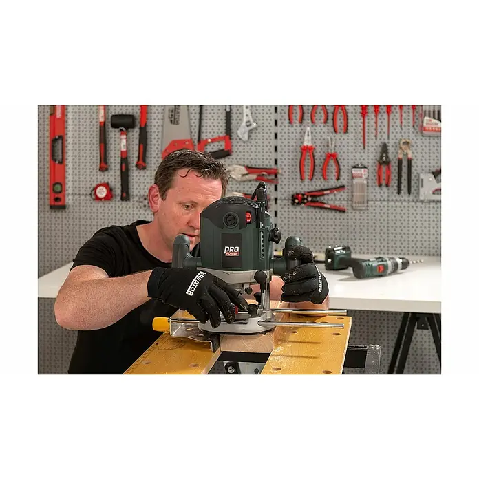 PowerPlus Fresadora eléctrica POWP6050 - 2.100 W, 8.000 r.p.m. - 23.500 r.p.m., Diámetro del portaherramientas: 12 mm - Un hombre opera una fresadora vertical PRO POWER en un banco de trabajo, dando forma a la madera. Herramientas colgadas en la pared al fondo.