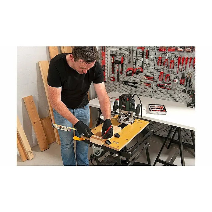 PowerPlus Fresadora eléctrica POWP6050 - 2.100 W, 8.000 r.p.m. - 23.500 r.p.m., Diámetro del portaherramientas: 12 mm - Hombre en taller con enrutador. Está marcando un trozo de madera con un lápiz mientras lo mide con una regla plegable. Herramientas en la pared.