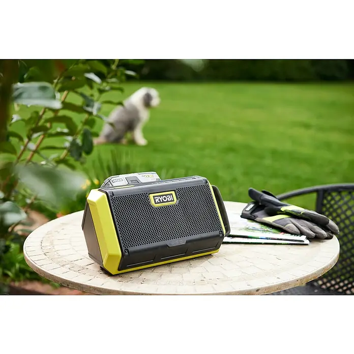 Ryobi ONE+ Altavoz Bluetooth RBT18-0 - 18 V, Sin batería, Potencia de salida altavoz: 15 W - Primer plano de un altavoz RYOBI ONE+ en una mesa de jardín, con guantes y un perro borroso al fondo.