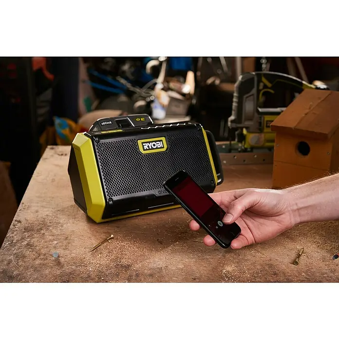 Ryobi ONE+ Altavoz Bluetooth RBT18-0 - 18 V, Sin batería, Potencia de salida altavoz: 15 W - Una radio Ryobi ONE+, un teléfono móvil y unos tornillos están sobre un banco de trabajo. La radio es amarilla y negra.