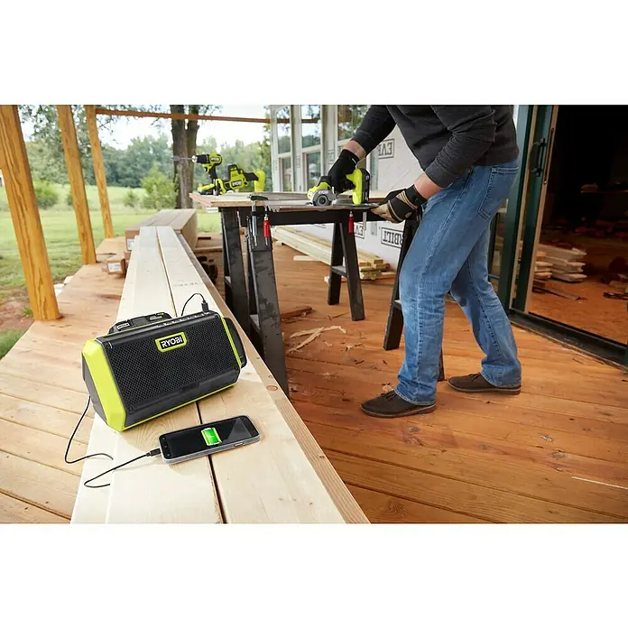 Ryobi ONE+ Altavoz Bluetooth RBT18-0 - 18 V, Sin batería, Potencia de salida altavoz: 15 W - Radio de obra Ryobi conectada a un teléfono inteligente, mientras un trabajador de la construcción trabaja con herramientas al fondo.