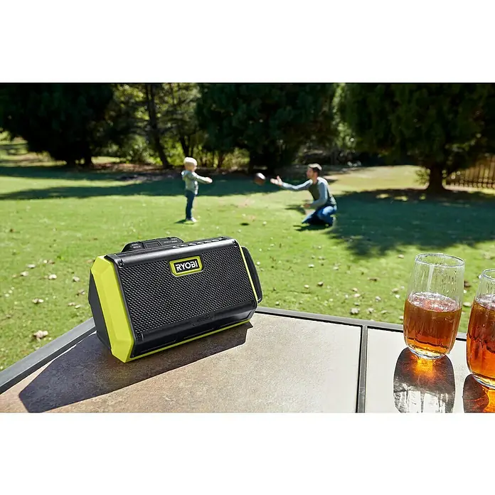 Ryobi ONE+ Altavoz Bluetooth RBT18-0 - 18 V, Sin batería, Potencia de salida altavoz: 15 W - Un altavoz Ryobi portátil negro y amarillo neón está sobre una mesa con bebidas mientras una familia juega al fondo.