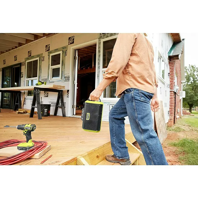 Ryobi ONE+ Altavoz Bluetooth RBT18-0 - 18 V, Sin batería, Potencia de salida altavoz: 15 W - Un artesano lleva una caja portátil Ryobi en una obra en construcción. Se destaca la construcción robusta y la movilidad del dispositivo.