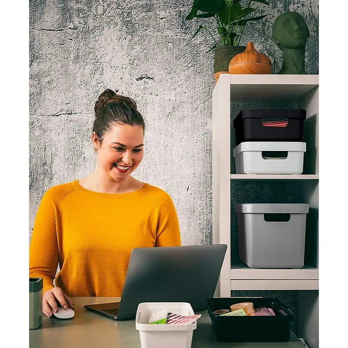 Sunware Aufbewahrungsbox Sigma Home - L x B x H: 35 x 24,6 x 12,2 cm, Kunststoff, Schwarz - Eine Frau sitzt am Schreibtisch und arbeitet am Laptop. Im Regal sind verschiedenfarbige Aufbewahrungsboxen zu sehen, die für Ordnung sorgen.