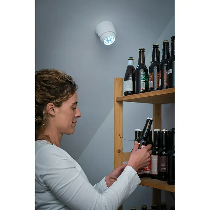 LED-Bewegungsmelder Vigilamp - Weiß, Außen - Frau wählt eine Bierflasche aus einem Regal unter einer batteriebetriebenen LED-Leuchte. Praktische und energiesparende Beleuchtung.