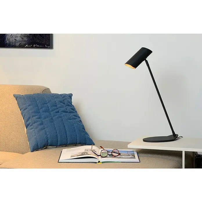 Lucide Tischleuchte Hester - 7 W, L x B x H: 12 x 21,5 x 54,5 cm, Anthrazit, GU10 - Eine elegante, schwarze Schreibtischlampe beleuchtet ein offenes Buch mit Brille darauf, neben einem blauen Kissen auf einem beigen Sofa.