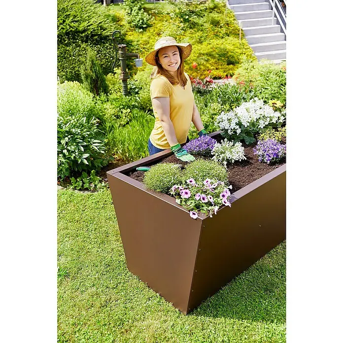 Gutta Hochbeet Valenta 150 - 150 x 90 x 80 cm, Aluminium, Rost Matt - Eine Frau in einem Garten arbeitet an einem großen, rechteckigen, braunen Pflanzgefäß mit blühenden Blumen.